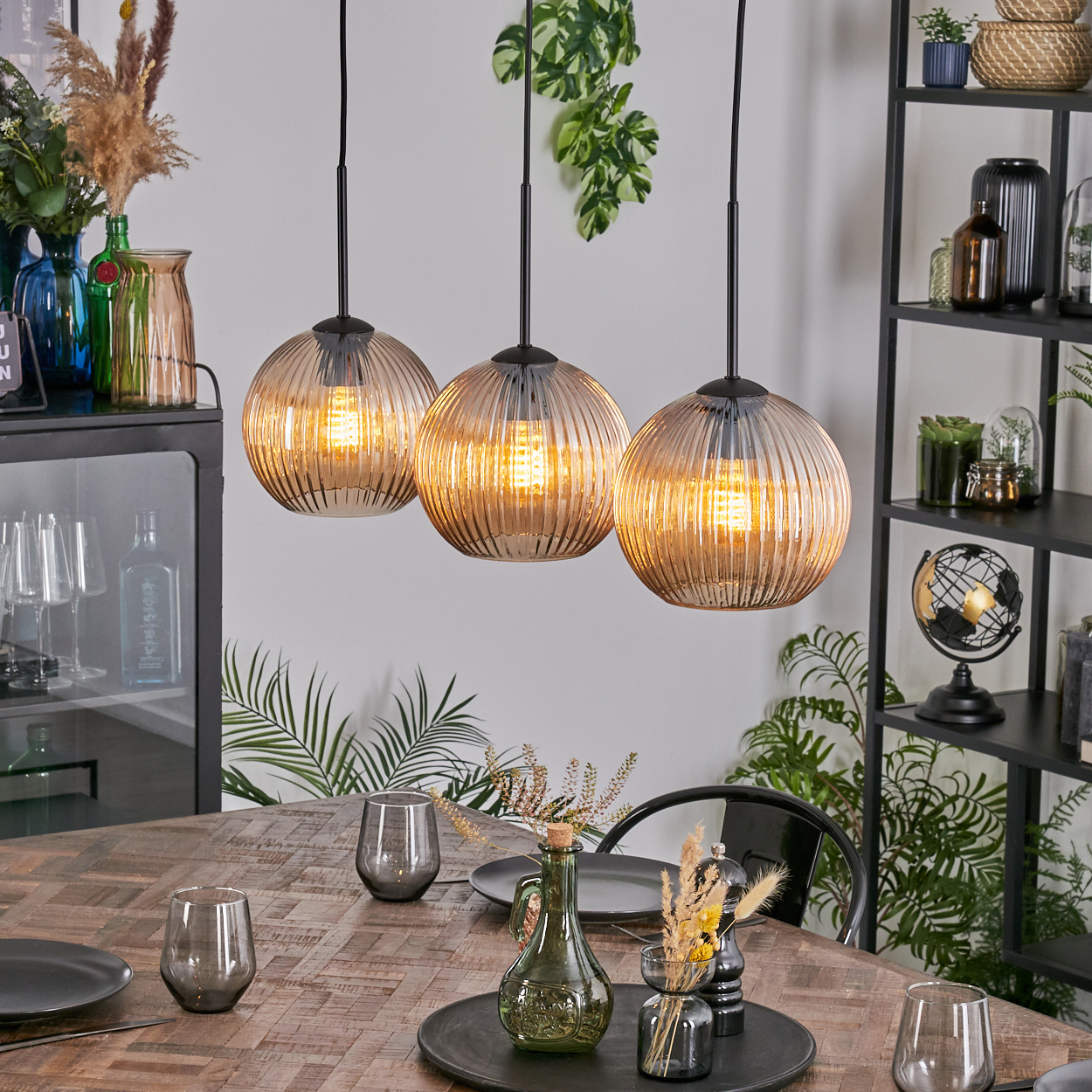 Esszimmer mit drei Hängelampen. Ein Holztisch ist mit Tellern und Gläsern gedeckt. Regale beherbergen Dekor, Pflanzen und einen Globus.