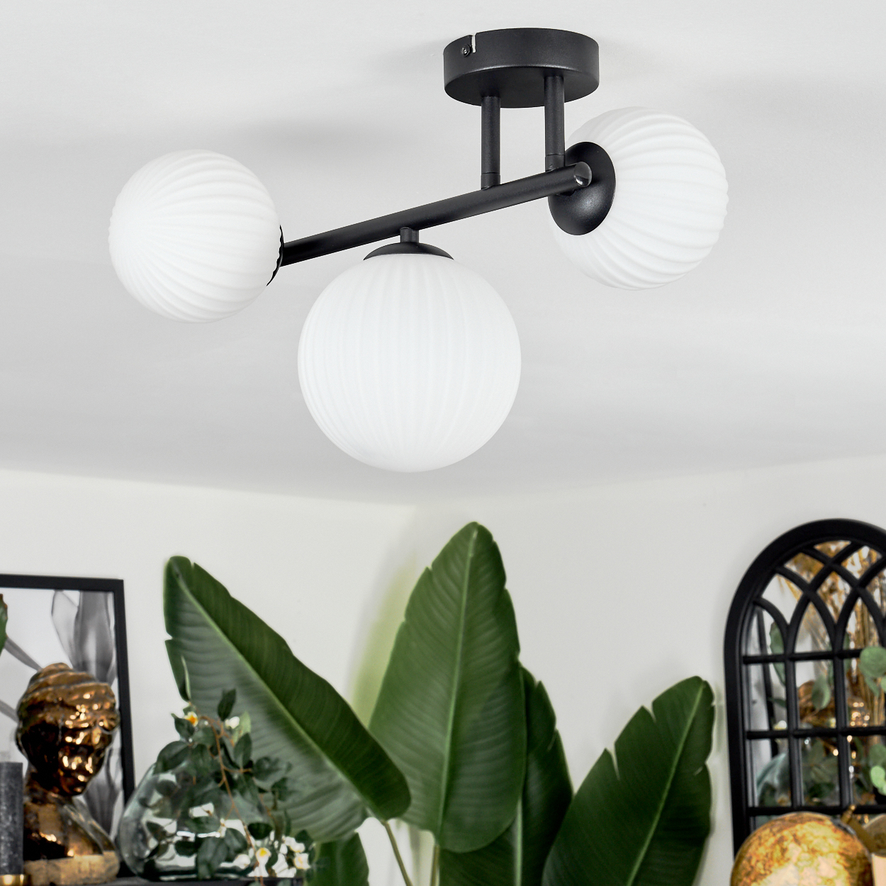 Eine moderne Deckenlampe mit drei weißen, kugelförmigen Lichtern. Sie hat einen schwarzen Metallrahmen. Grüne Pflanzen im Vordergrund.