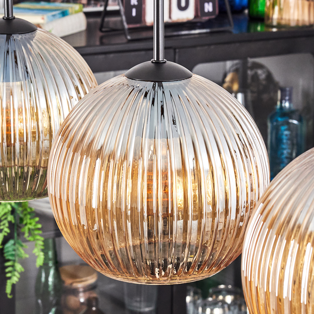 Drei kugelförmige, gerippte Glas-Pendelleuchten mit warmem Innenlicht. Der Hintergrund umfasst ein Regal mit Flaschen und Pflanzen.