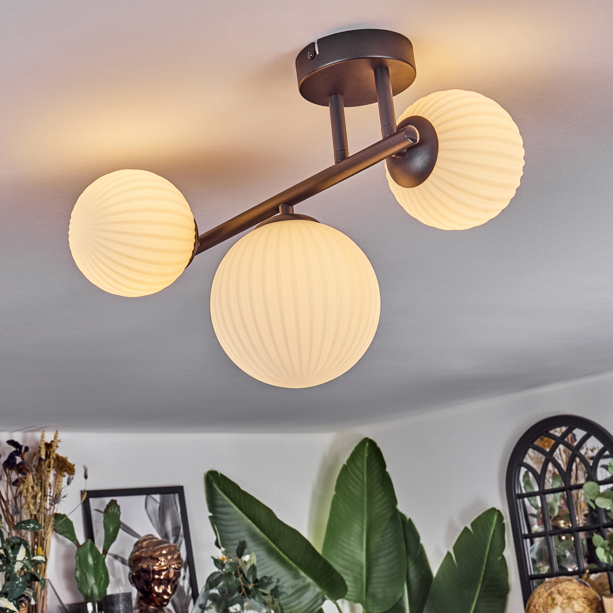Eine moderne Deckenleuchte mit drei kugelförmigen Lampen. Die Lampen sind cremefarben und gerippt. Grüne Pflanzen und Dekorationen im Hintergrund.