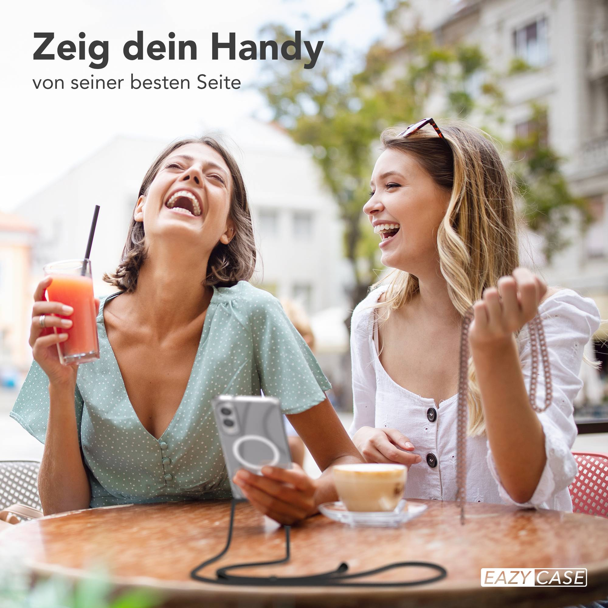 Zwei lachende Frauen im Freien. Eine hält ein Getränk, die andere eine Halskette und ein Telefon mit Hülle.