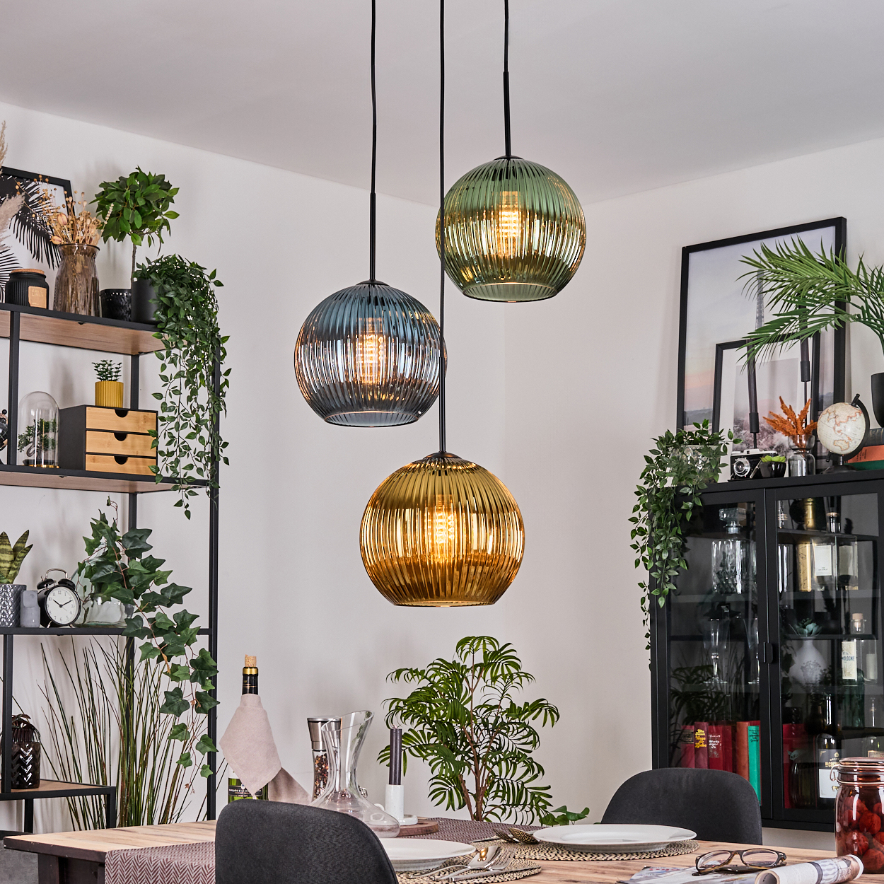 Drei Pendelleuchten mit Glaskugeln hängen über einem Tisch. Der Raum hat Pflanzen, Regale und einen Schrank mit Dekorationsartikeln.