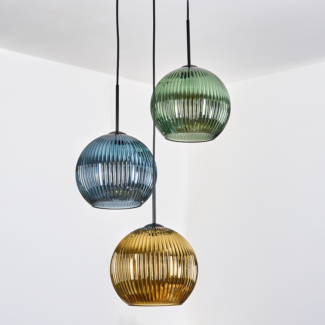 Drei kugelförmige Pendelleuchten in Blau, Grün und Gold hängen von der Decke. Der Hintergrund ist eine weiße Ecke.