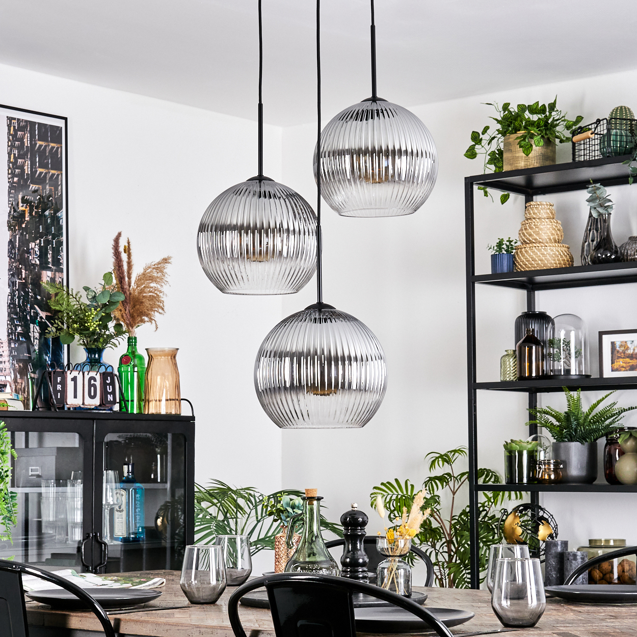Esszimmer mit drei Hängelampen. Tisch gedeckt, Pflanzen und ein schwarzes Regal im Hintergrund zu sehen.