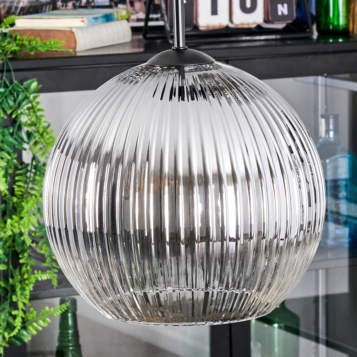 Eine klare, gerippte Glas-Pendelleuchte hängt von der Decke. Dahinter ein Regal mit Büchern, Pflanzen und Dekorationsartikeln.