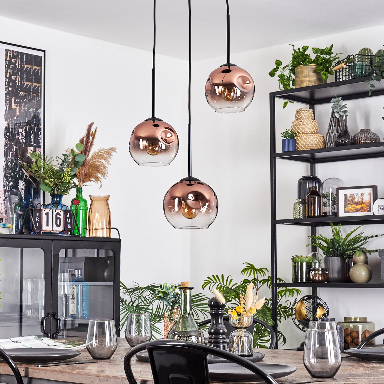Esszimmer mit kupferfarbenen Pendelleuchten, Holztisch, schwarzen Stühlen und einem Metallregal mit Pflanzen.
