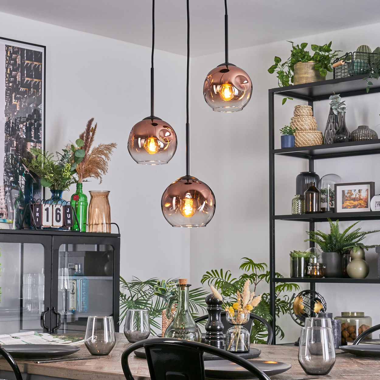 Esszimmer mit drei Pendelleuchten. Tisch mit Gläsern und Tellern gedeckt. Regale mit Pflanzen und Dekoration.