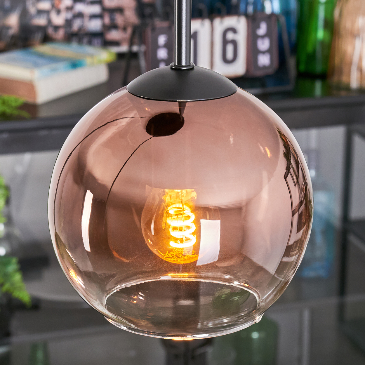 Eine Pendelleuchte aus Bronzeglas mit einer Wendelglühbirne. Im Hintergrund ein Kalender, Bücher und grüne Flaschen.