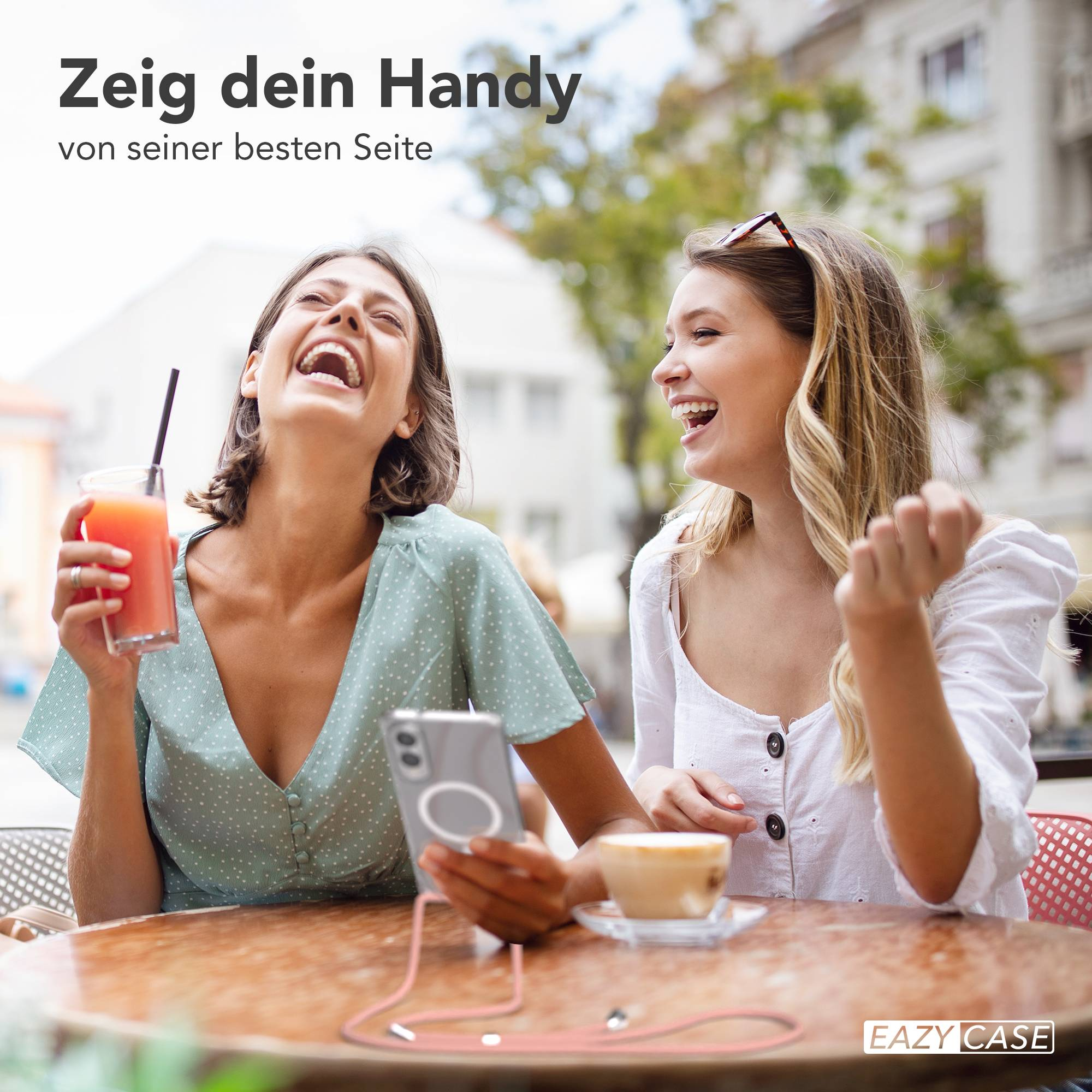 Zwei lachende Frauen in einem Café, eine hält ein Getränk, die andere eine Sonnenbrille. Ein Telefon mit Hülle ist sichtbar.