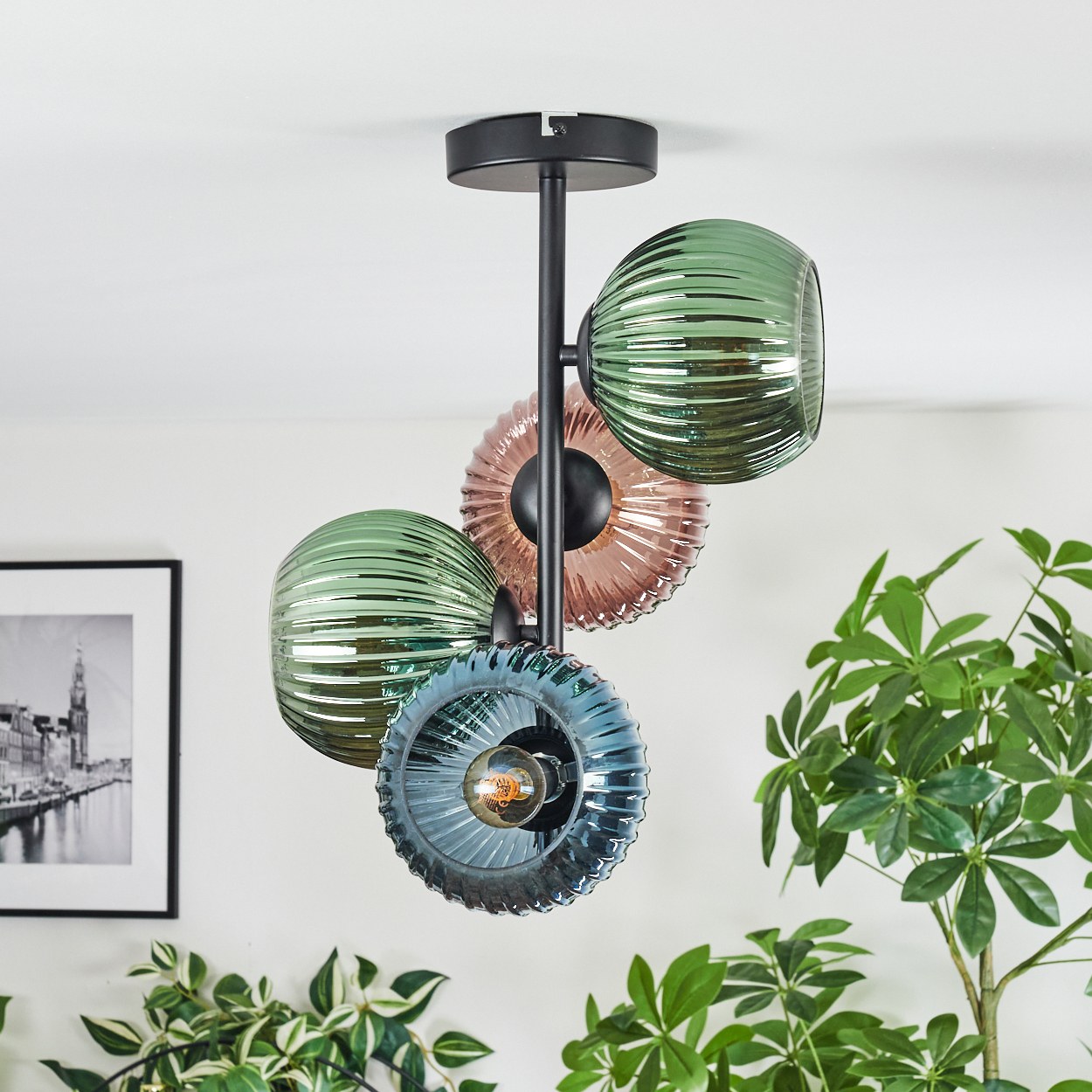 Eine moderne Deckenlampe mit grünen, rosa und blauen Glasschirmen. Grüne Pflanzen und ein gerahmtes Bild sind im Hintergrund.