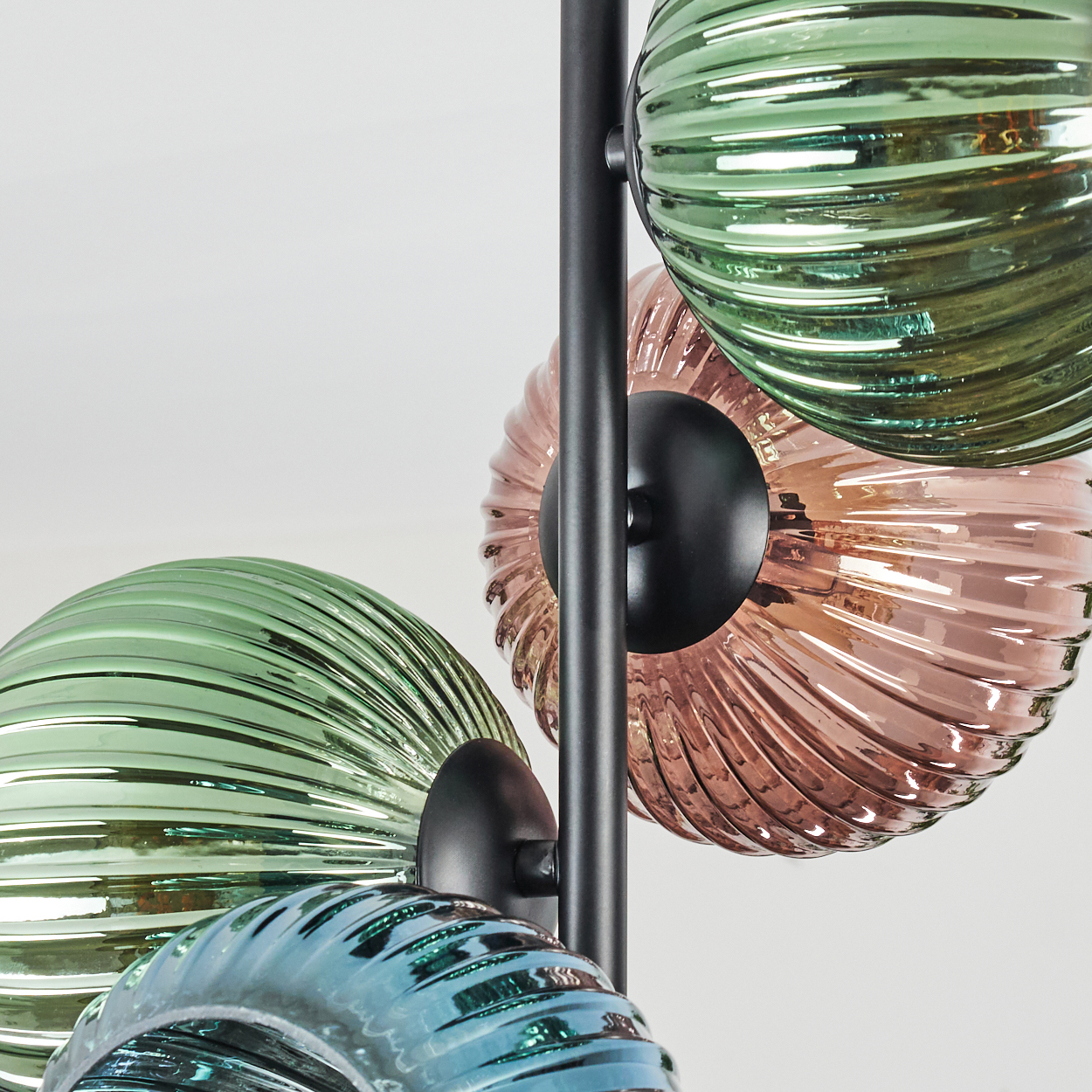 Nahaufnahme einer Lampe mit gerippten, farbigen Glaskugeln. Farben sind Grün, Blau und Rosa. Schwarze Metallstütze. Weißer Hintergrund.