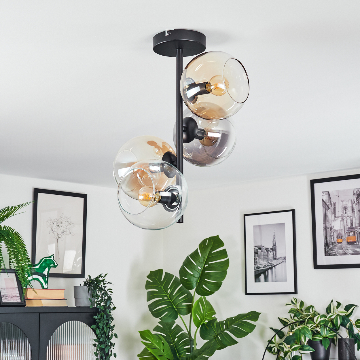 Eine moderne Deckenlampe mit Glaskugeln hängt von einer weißen Decke. Darunter schmücken eine grüne Pflanze und gerahmte Bilder einen Raum.