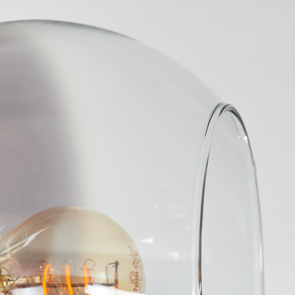 Nahaufnahme eines gläsernen Lampenschirms mit sichtbarer Glühbirne. Das Glas ist klar und die Glühbirne hat orangefarbene Filamente. Der Hintergrund ist weiß.