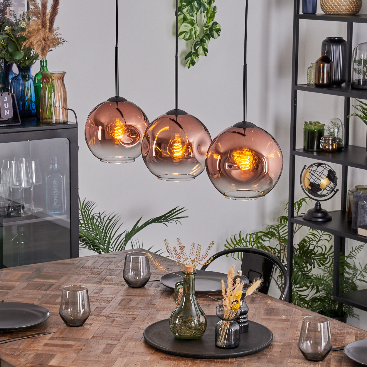 Drei kupferfarbene Pendelleuchten hängen über einem Holztisch, der mit Gläsern und Tellern gedeckt ist. Im Hintergrund sind Regale und Pflanzen zu sehen.