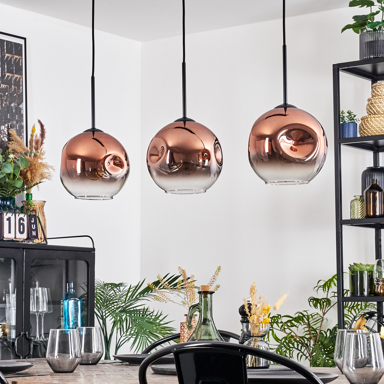 Drei kupferfarbene Pendelleuchten hängen über einem Tisch. Die Szene umfasst Pflanzen, Gläser und ein schwarzes Metallregal mit Dekoration.