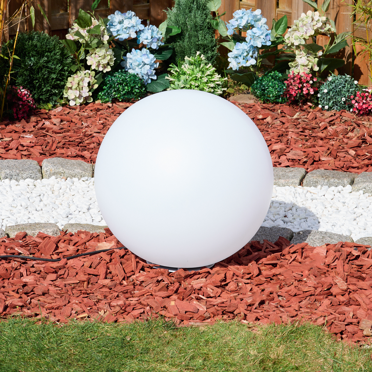 Eine große weiße Kugel liegt in einem Gartenbeet mit roter Mulch, weißen Steinen und Blumen. Grünes Gras ist im Vordergrund.