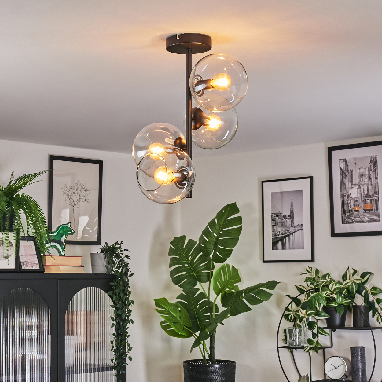 Ein modernes Wohnzimmer mit Kronleuchter, Pflanzen und gerahmten Bildern. Ein schwarzer Schrank und ein dekoratives Metallregal sind ebenfalls sichtbar.