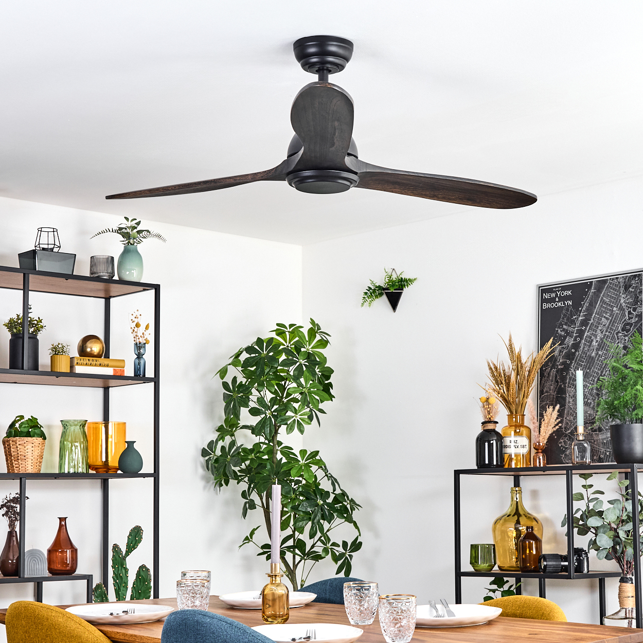 Ein dunkler Deckenventilator aus Holz hängt über einem für das Abendessen gedeckten Esstisch. Regale mit Vasen und Pflanzen flankieren den Raum.