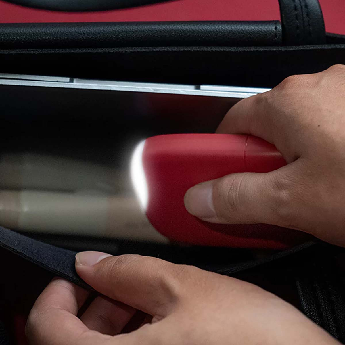 Hände halten ein rotes, rechteckiges Objekt mit weißem Licht in der Nähe eines Laptops in einer schwarzen Tasche. Rote und schwarze Details sind sichtbar.