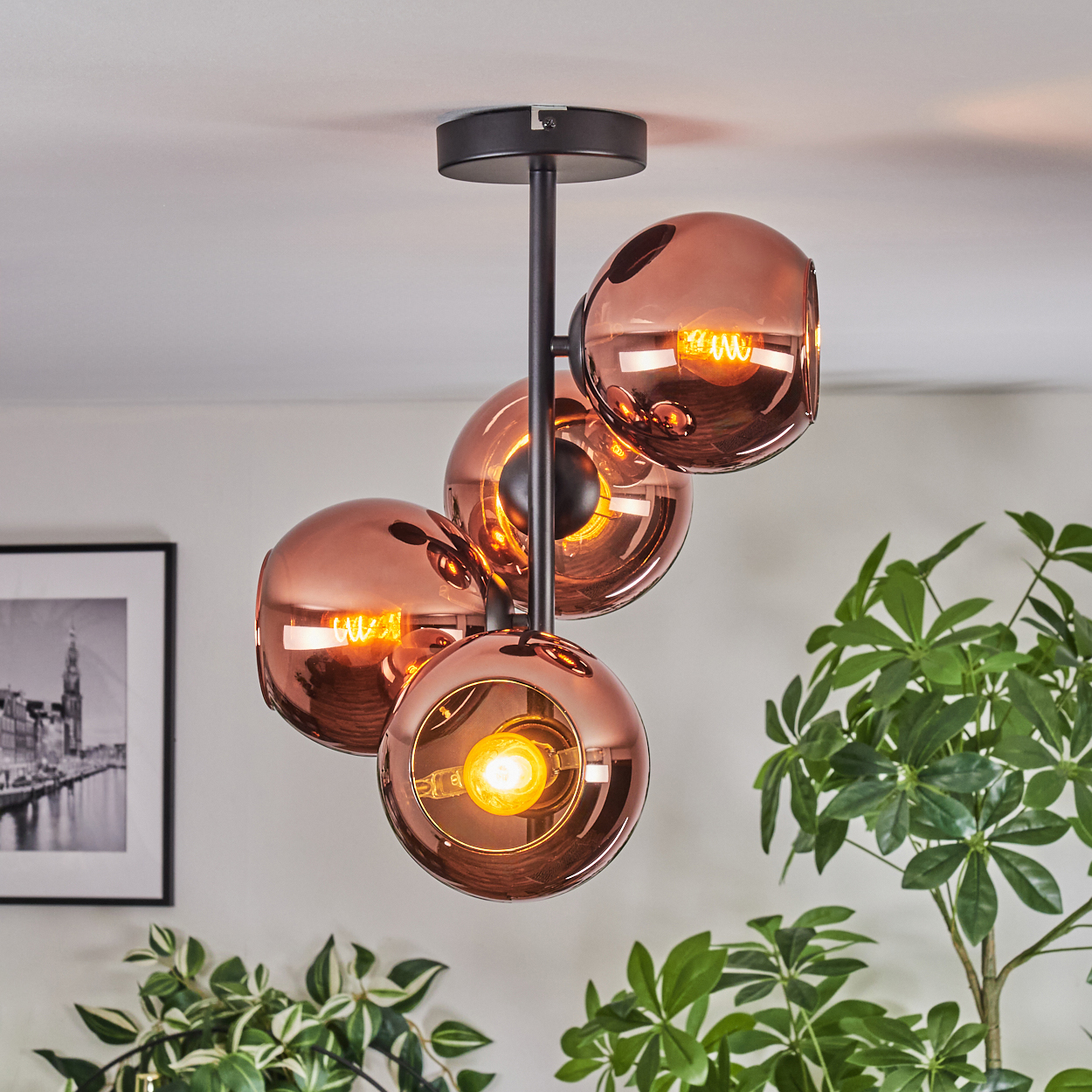 Eine moderne Deckenlampe mit vier kupferfarbenen Glaskugeln, jede mit einer warmen Glühbirne. Im Hintergrund grüne Pflanzen.