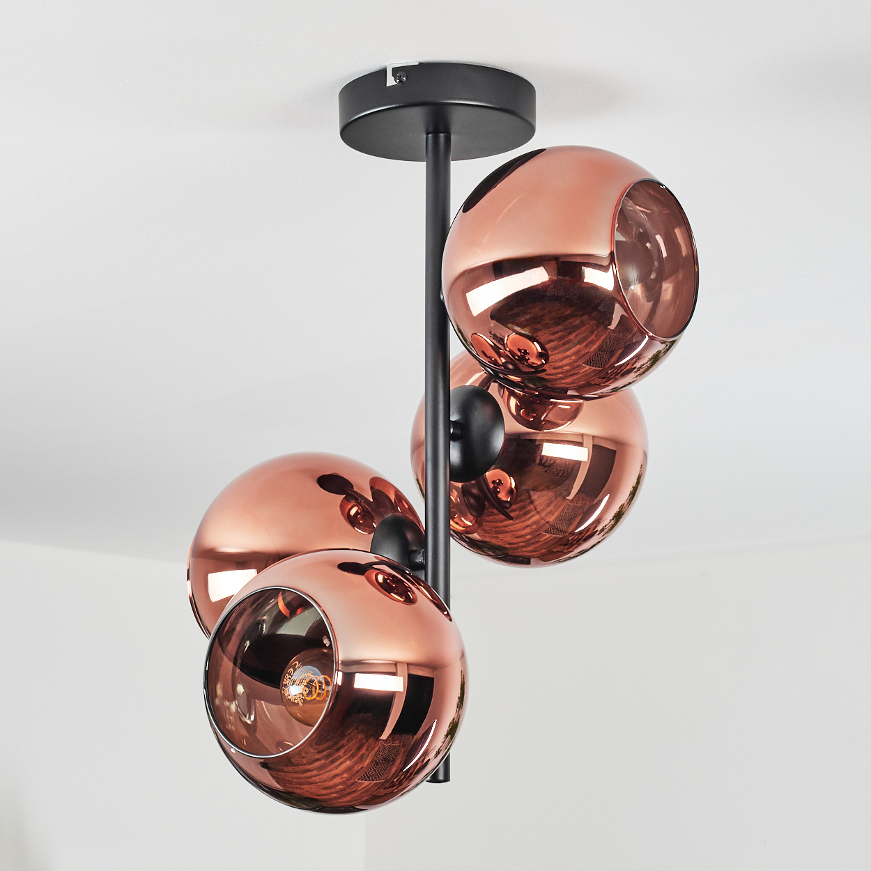 Eine moderne Deckenleuchte mit vier kupferfarbenen Kugelschirmen, die an einem schwarzen Metallrahmen hängen, vor einer weißen Decke.