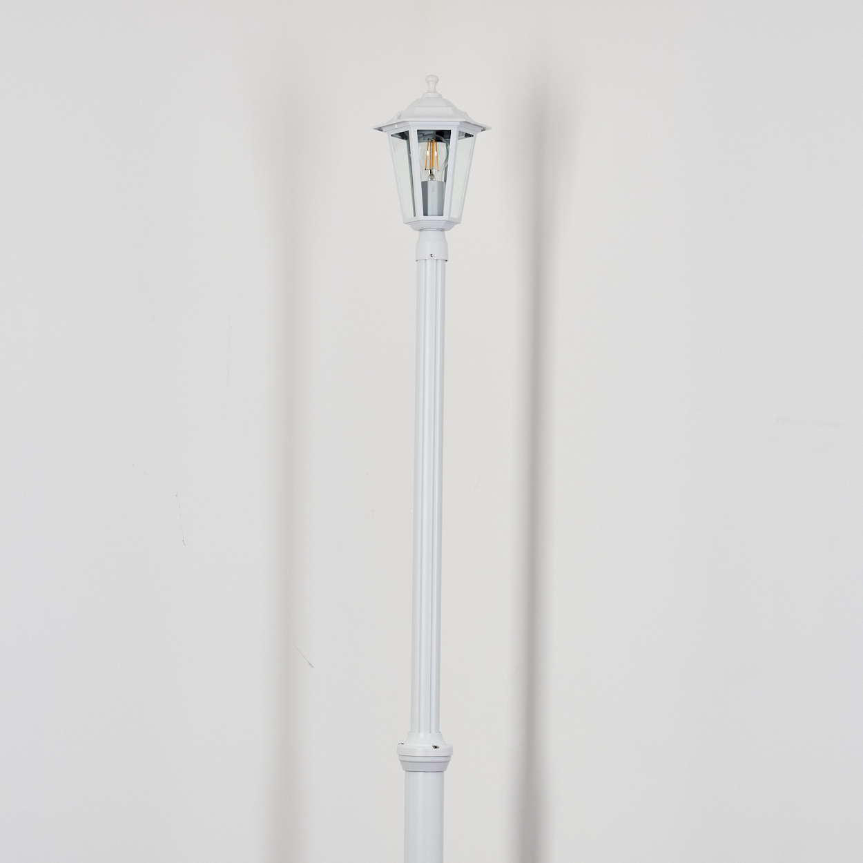 Eine weiße Laterne steht vor einer weißen Wand. Die Laterne hat ein Glasgehäuse und eine Glühbirne. Ein Schatten wird auf die Wand geworfen.