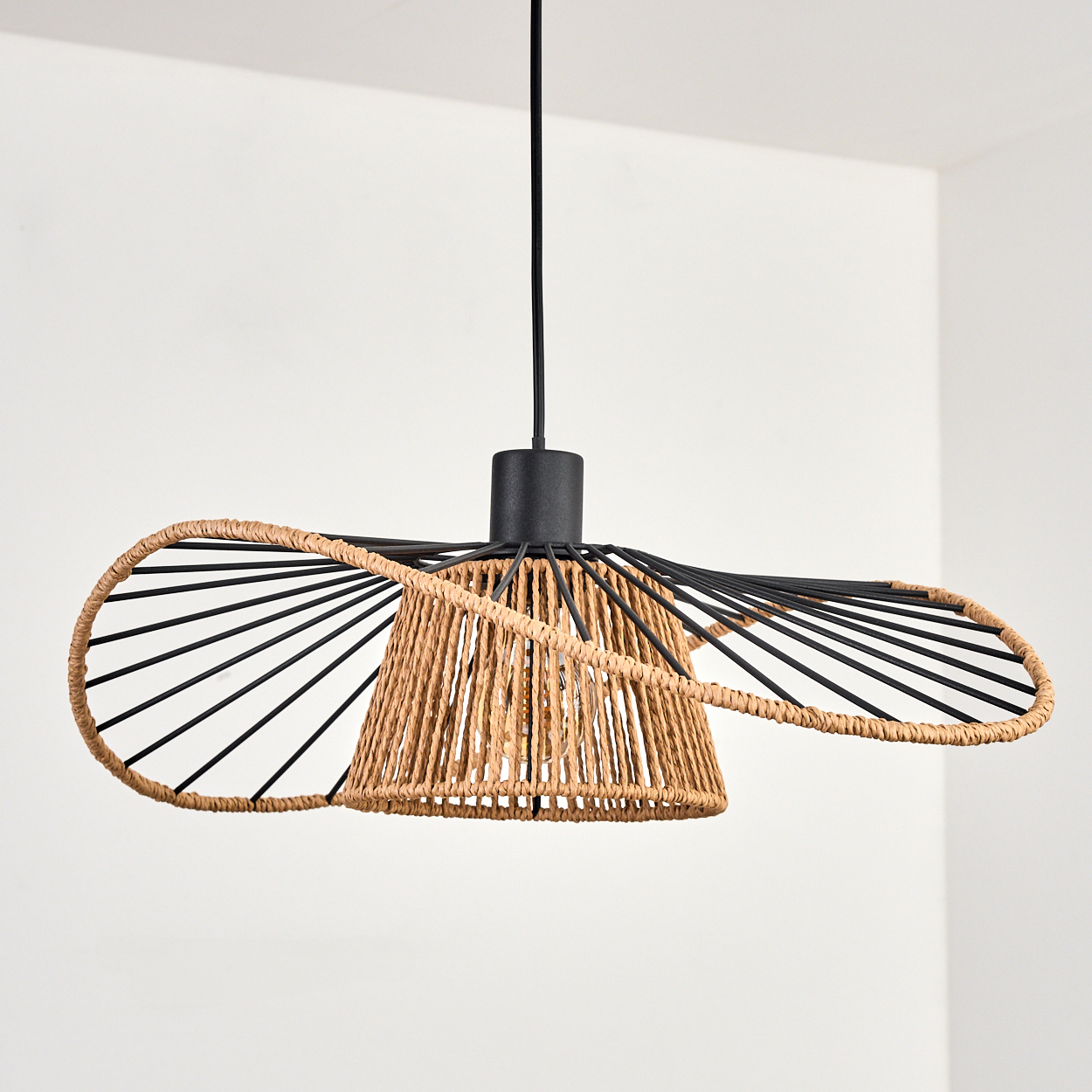 Eine moderne Pendelleuchte mit gewebtem Lampenschirm und schwarzem Metallrahmen, vor einer weißen Wand hängend.