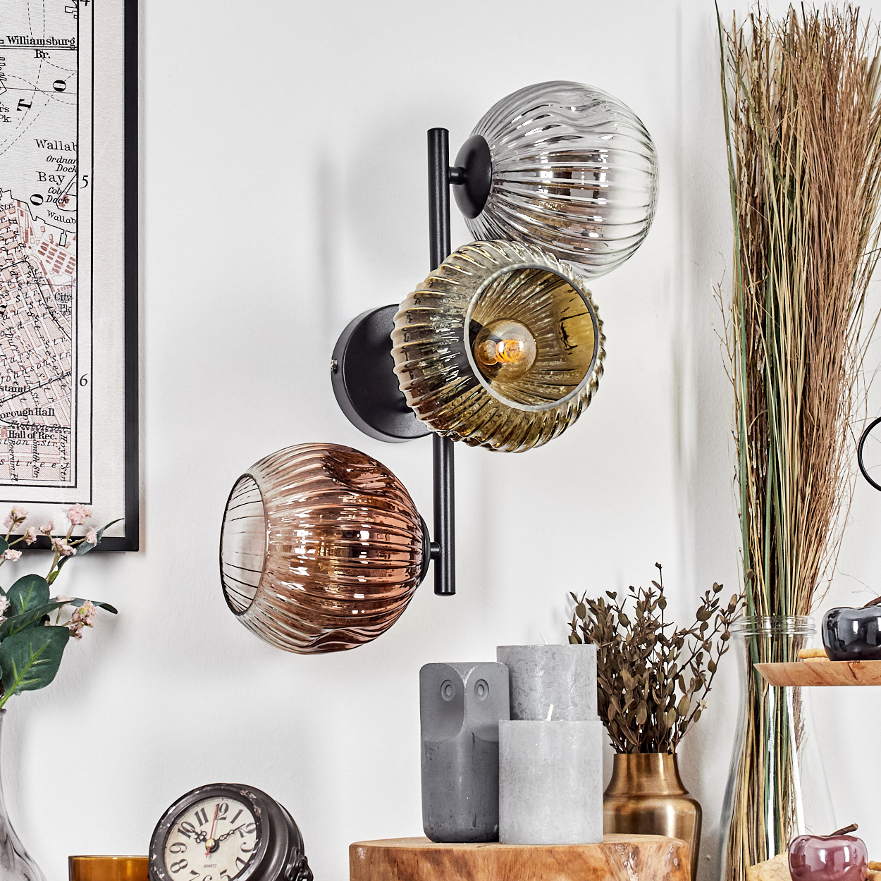 Eine Wandlampe mit drei Glaskugeln, eine Uhr, Kerzen und Dekorationsartikel an einer weißen Wand.