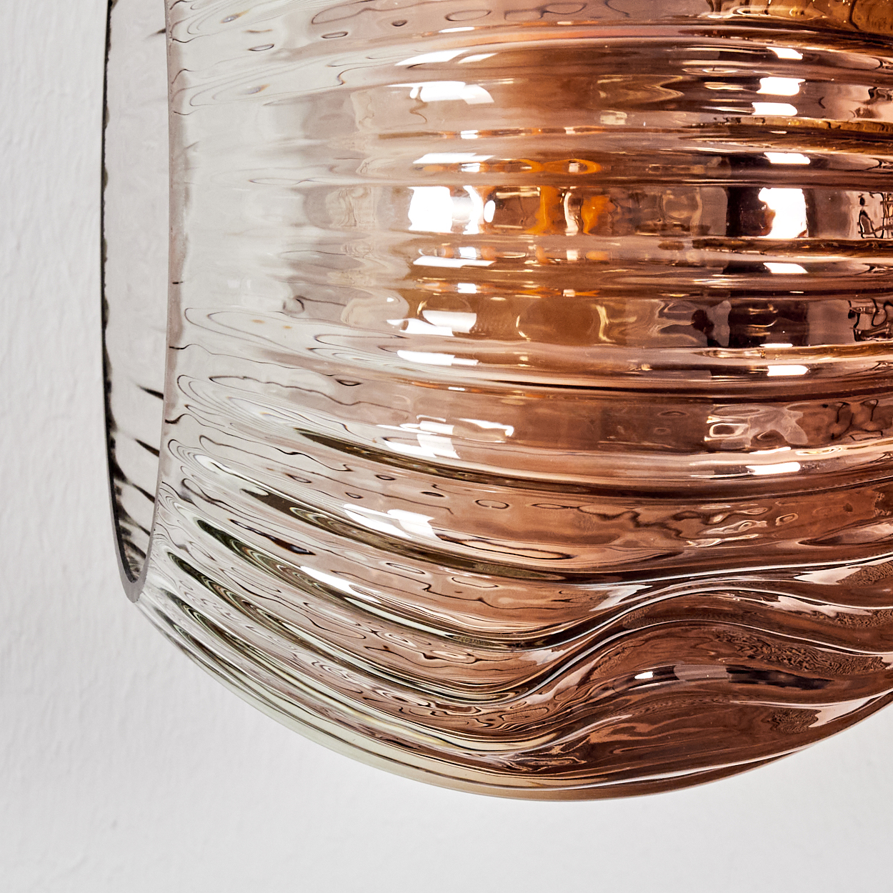Nahaufnahme eines geschwungenen Glasobjekts mit wellenförmigen Linien. Es hat einen Bronzeton und reflektiert Licht. Der Hintergrund ist eine weiße Wand.