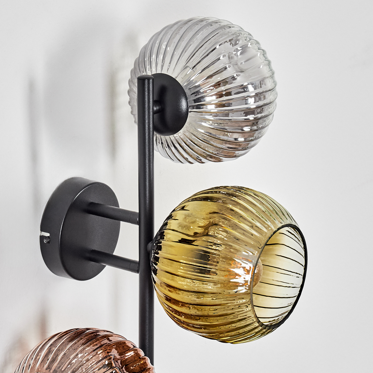 Eine moderne Wandlampe mit drei kugelförmigen Glasschirmen in Silber-, Gelb- und Kupferfarben, montiert auf einem schwarzen Metallrahmen.