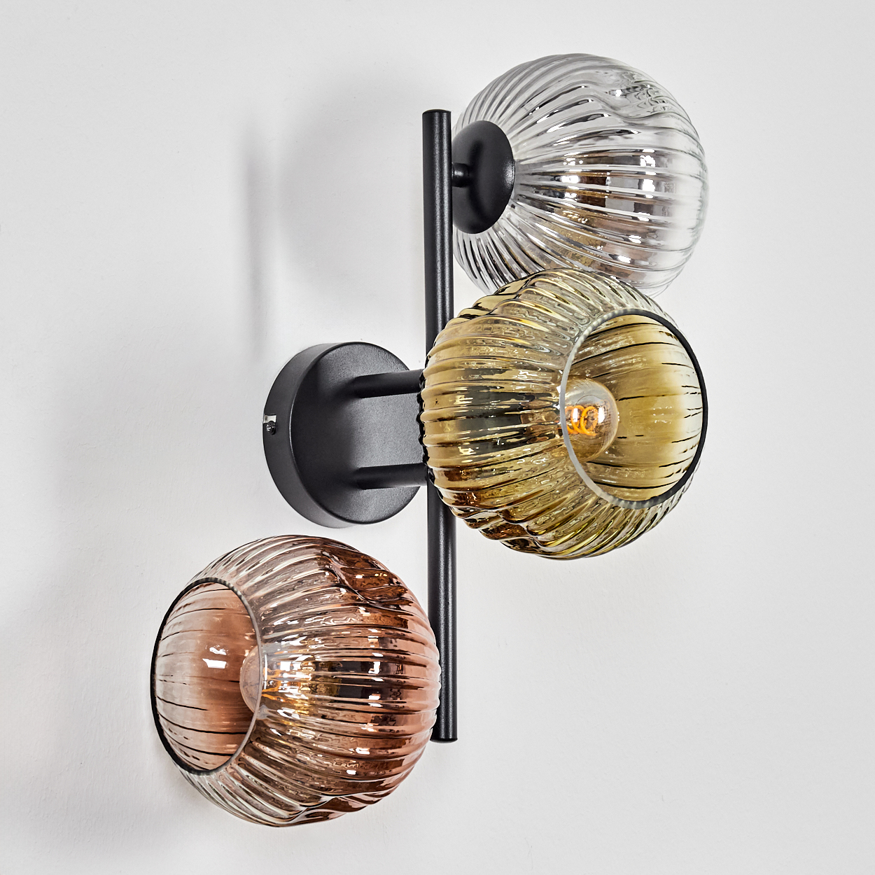 Eine moderne Wandlampe mit drei Glaskugeln in Silber, Bernstein und Kupfer, montiert auf einem schwarzen Metallrahmen, an einer weißen Wand.