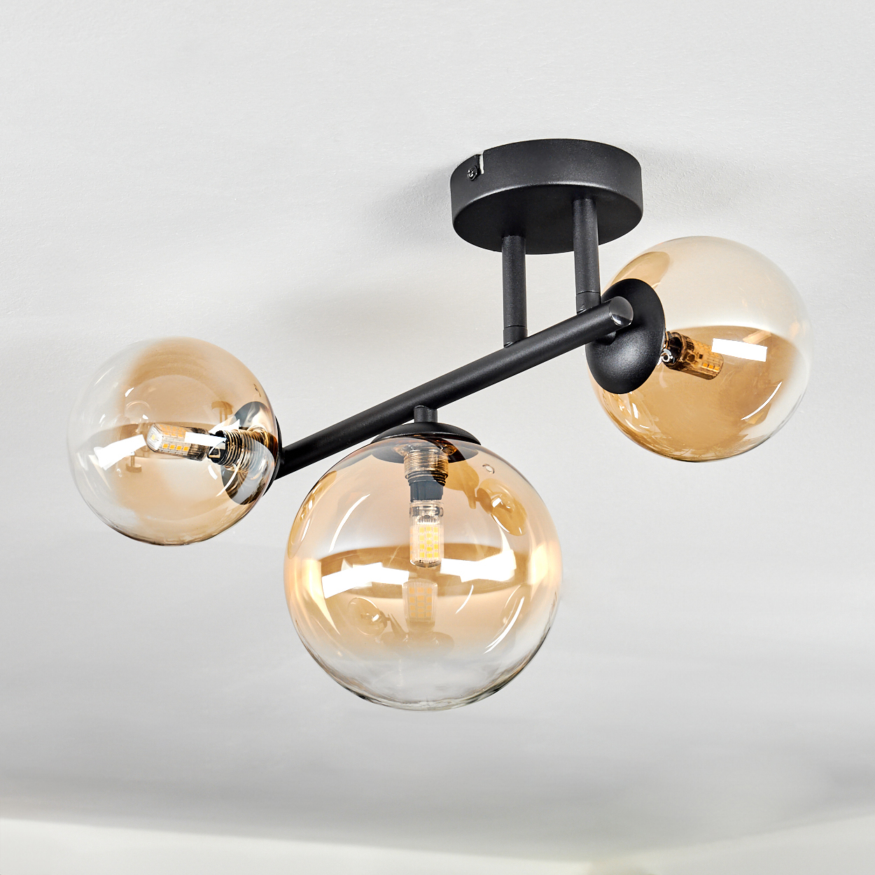 Eine moderne Deckenleuchte mit drei bernsteinfarbenen Glaskugeln. Schwarze Metallarme und -befestigung an der weißen Decke.