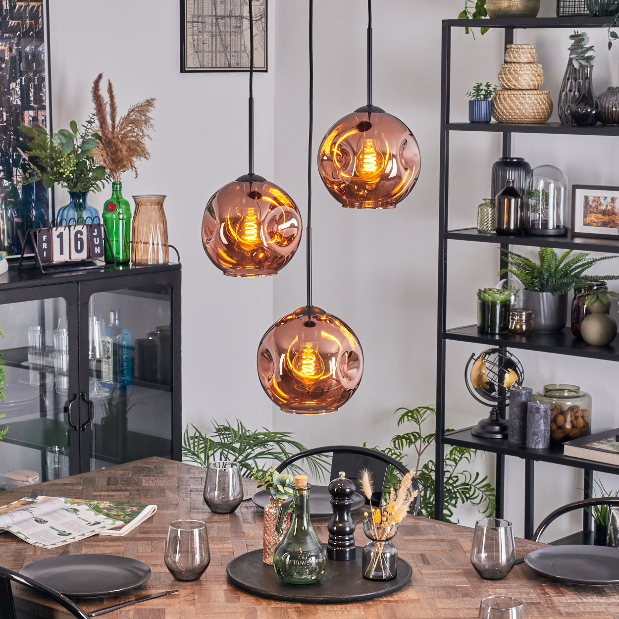 Esszimmer mit drei kupferfarbenen Pendelleuchten. Tisch mit Tellern und Gläsern. Regal mit Pflanzen und Deko.