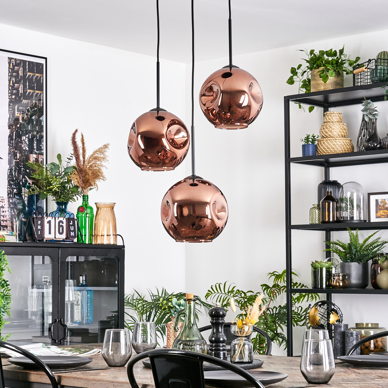 Esszimmer mit kupferfarbenen Pendelleuchten. Tisch mit Gläsern und Tellern gedeckt. Regale mit Pflanzen und Dekor im Hintergrund.