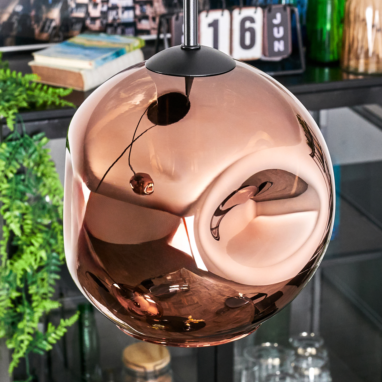 Eine kupferfarbene, kugelförmige Lampe hängt. Die Lampe reflektiert den Raum, mit Grün, Büchern und einem Kalender.