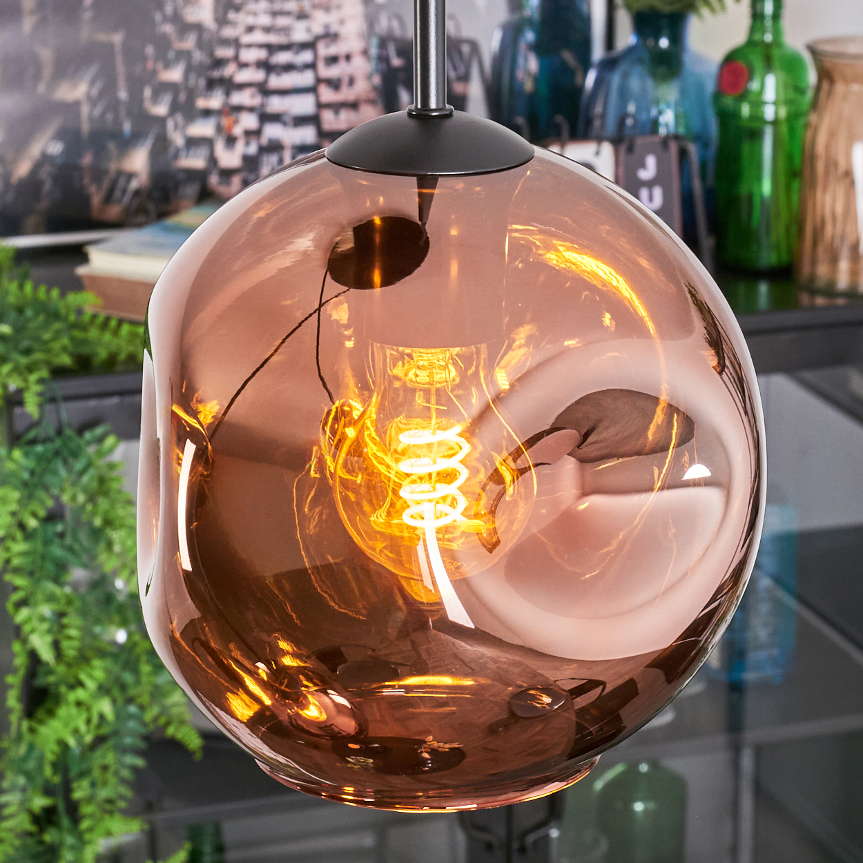 Kupferfarbene Glas-Hängeleuchte mit leuchtender Wendelglühbirne. Pflanzen und andere Dekorationen sind im Hintergrund zu sehen.
