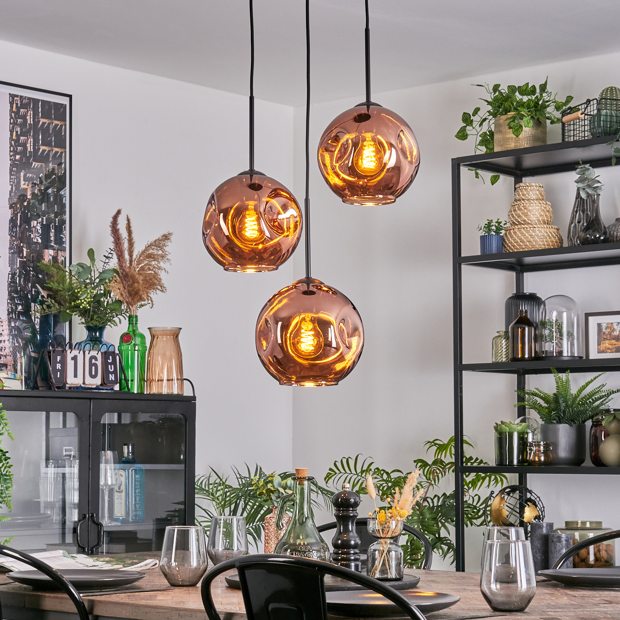 Esszimmer mit drei kupferfarbenen Pendelleuchten. Tisch mit Gläsern und Tellern gedeckt. Regale mit Pflanzen.