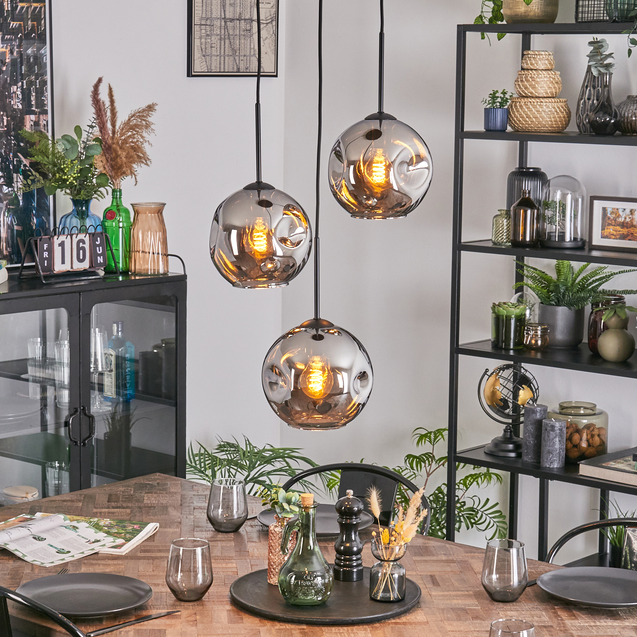 Esszimmer mit Holztisch, drei Hängelampen, schwarzes Regal mit Pflanzen und verschiedenen Dekorationen.