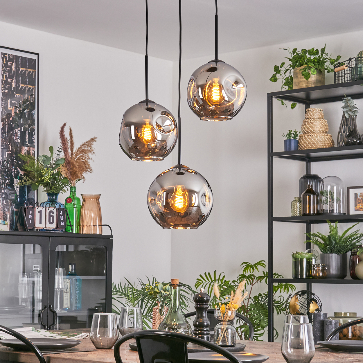 Esszimmer mit Hängelampen, Tischgedeck und einem schwarzen Regal mit Pflanzen und Dekorationen. Die Lampen haben leuchtende Glühbirnen.