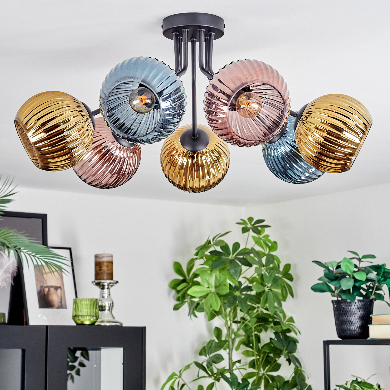 Moderne Deckenlampe mit sechs farbigen Glasschirmen. Die Farben sind Gold, Rosa und Blau. Pflanzen und Möbel sind im Hintergrund.