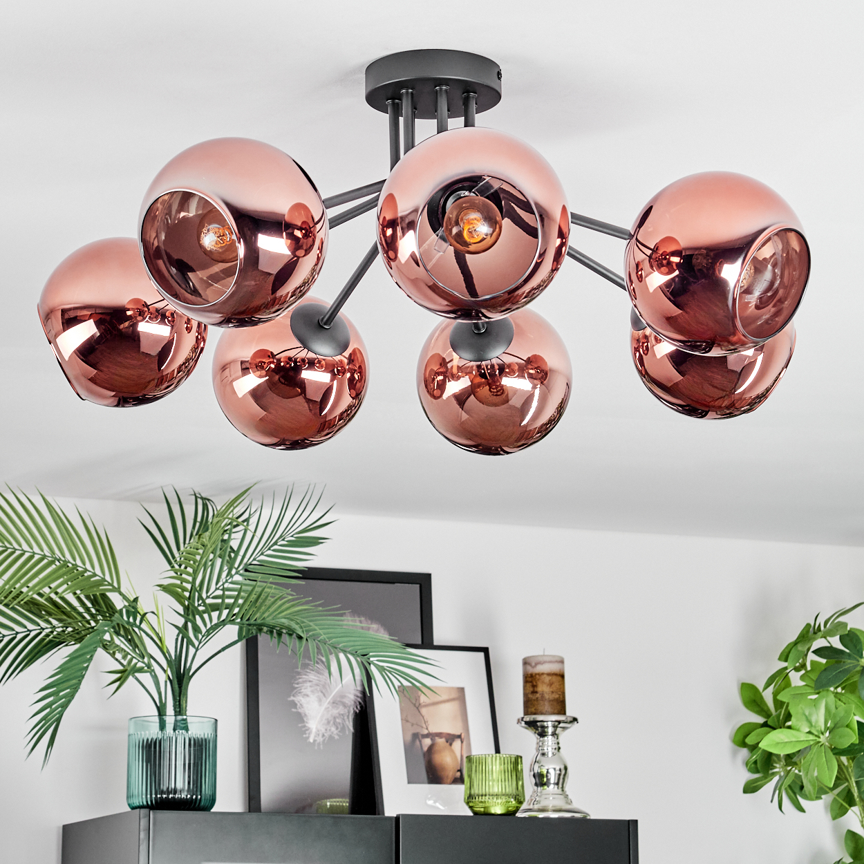 Eine moderne Deckenlampe mit kupferfarbenen Kugelschirmen. Darunter ein Tisch mit Pflanzen, Bildern und Kerzen.