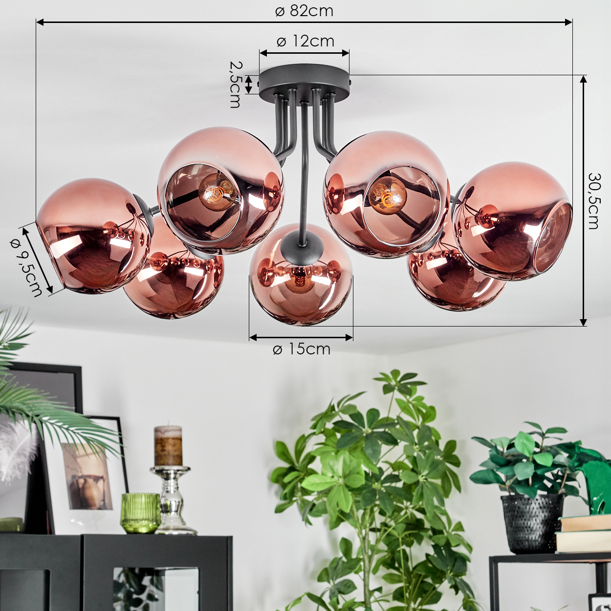 Eine moderne Deckenlampe mit sechs kupferfarbenen Kugelschirmen. Die Lampe ist an einer weißen Decke befestigt. Grüne Pflanzen sind im Vordergrund.