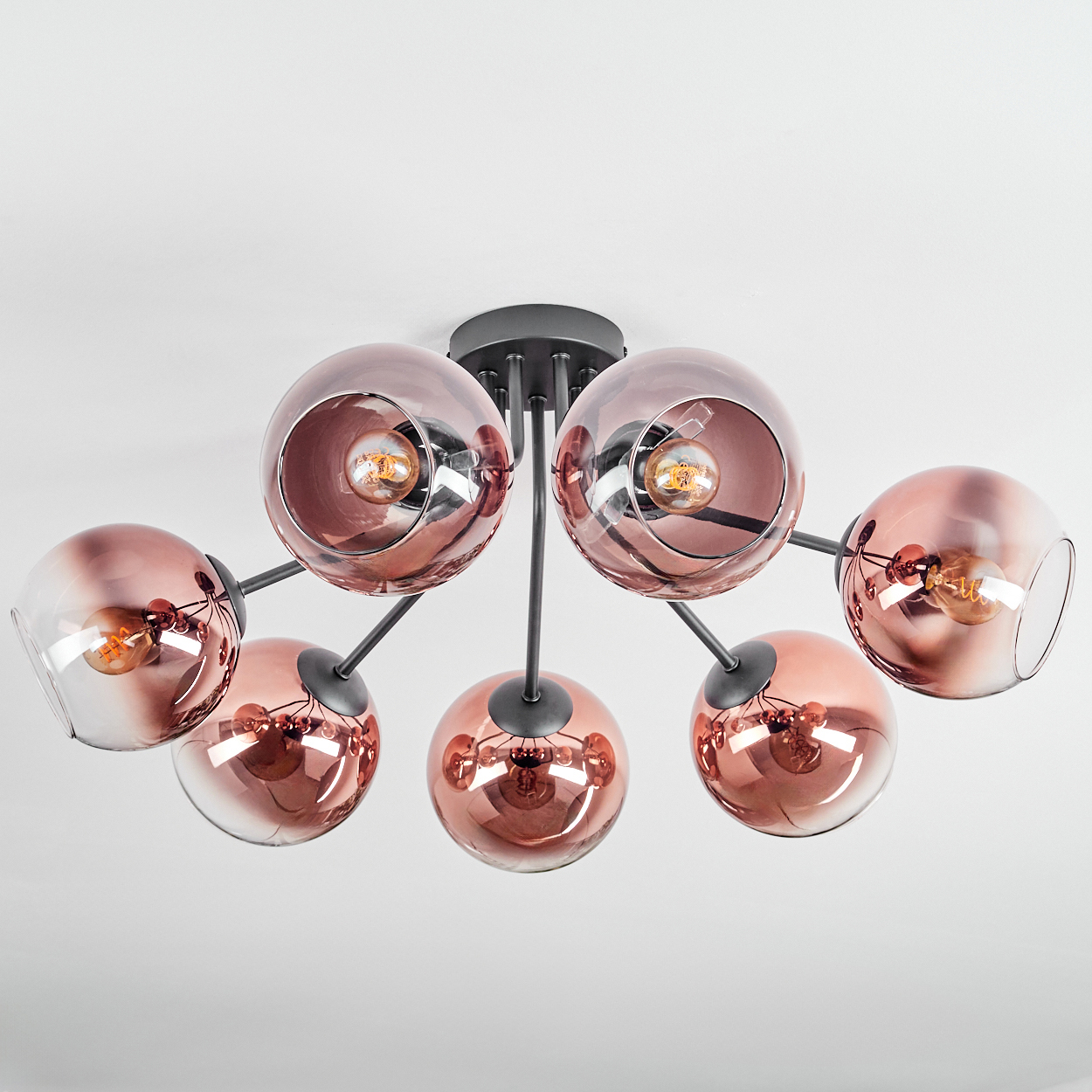 Eine moderne Deckenleuchte mit sieben kugelförmigen, roségoldfarben getönten Glasschirmen, jede mit einer Glühbirne, vor weißem Hintergrund.
