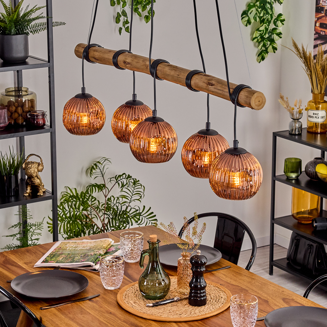 Esszimmer mit Holztisch, Kupfer-Hängelampen und Pflanzen. Tisch gedeckt mit Tellern, Gläsern und einer Flasche. Regale mit Dekoration.