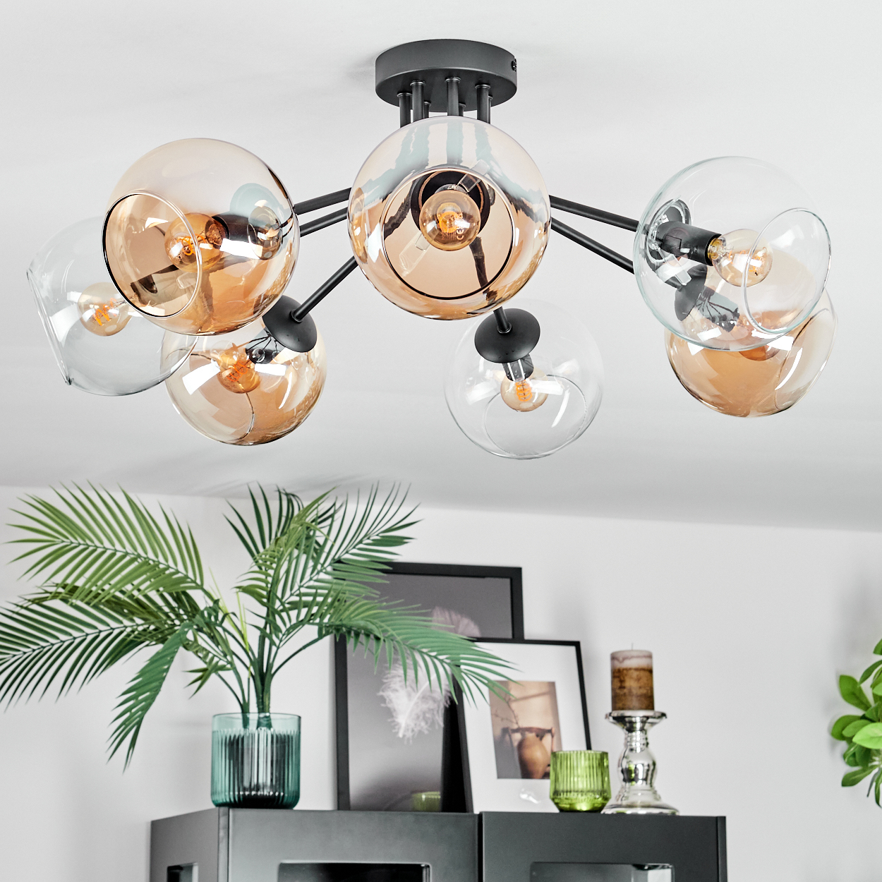 Eine moderne Deckenleuchte mit Glaskugeln hängt über einem Regal mit Dekoration, darunter Pflanzen, Bilder und Kerzen.