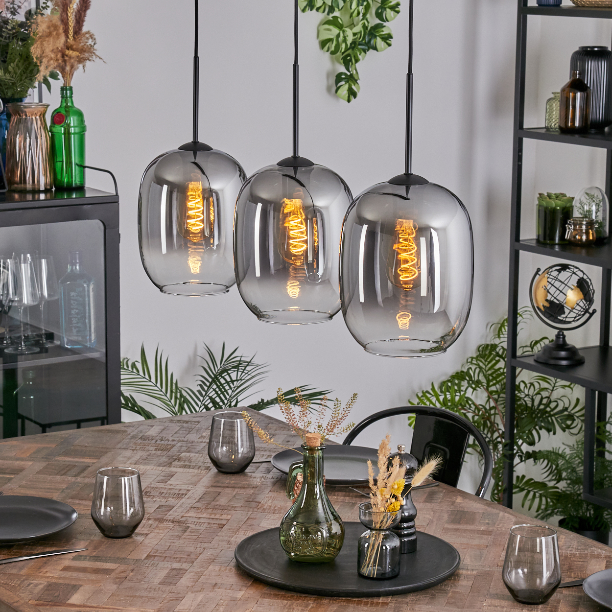 Drei Pendelleuchten hängen über einem Holztisch, der mit Gläsern und Tellern gedeckt ist. Regale und Schränke mit Deko sind im Hintergrund.