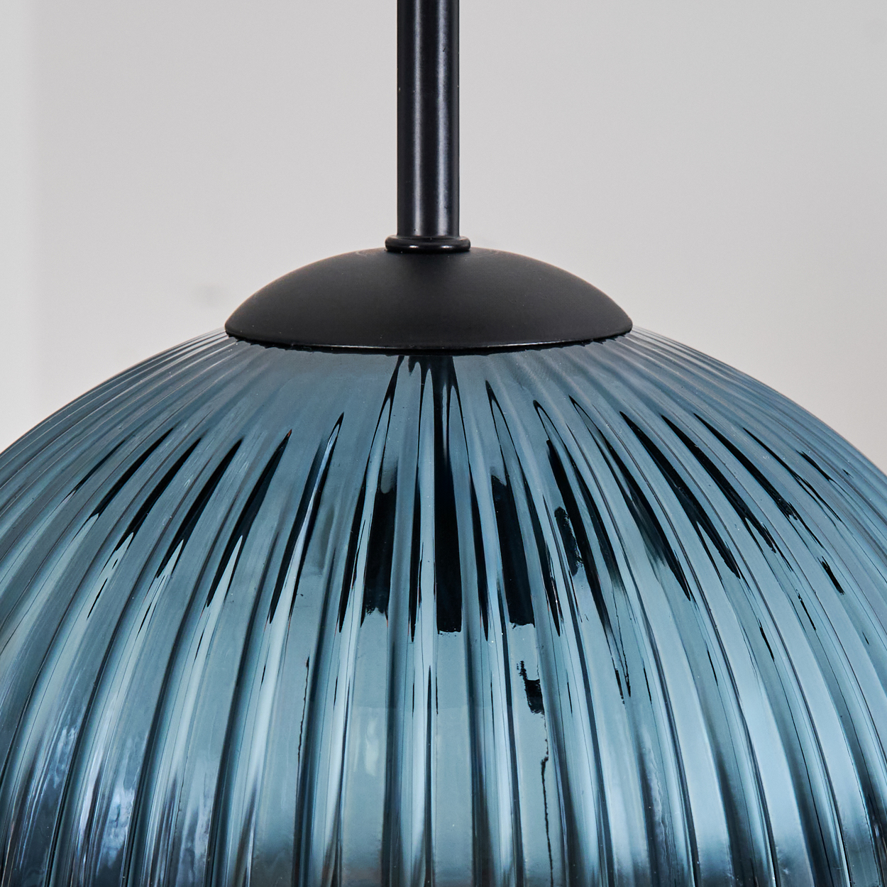 Nahaufnahme eines blauen Glaslampenschirms mit vertikalen Rippen, der an einer schwarzen Metallhalterung hängt, vor einer weißen Wand.