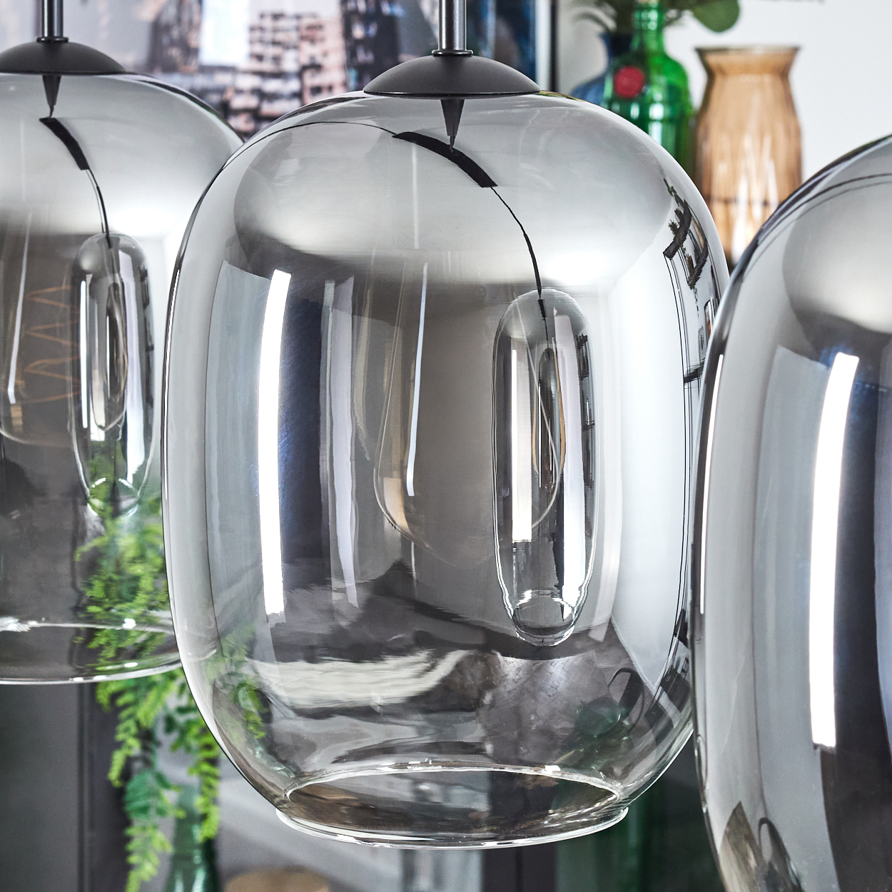 Nahaufnahme von drei modernen Glas-Pendelleuchten. Das Glas ist reflektierend, mit dunkler Deckenhalterung. Hintergrund: Vasen und Pflanzen.