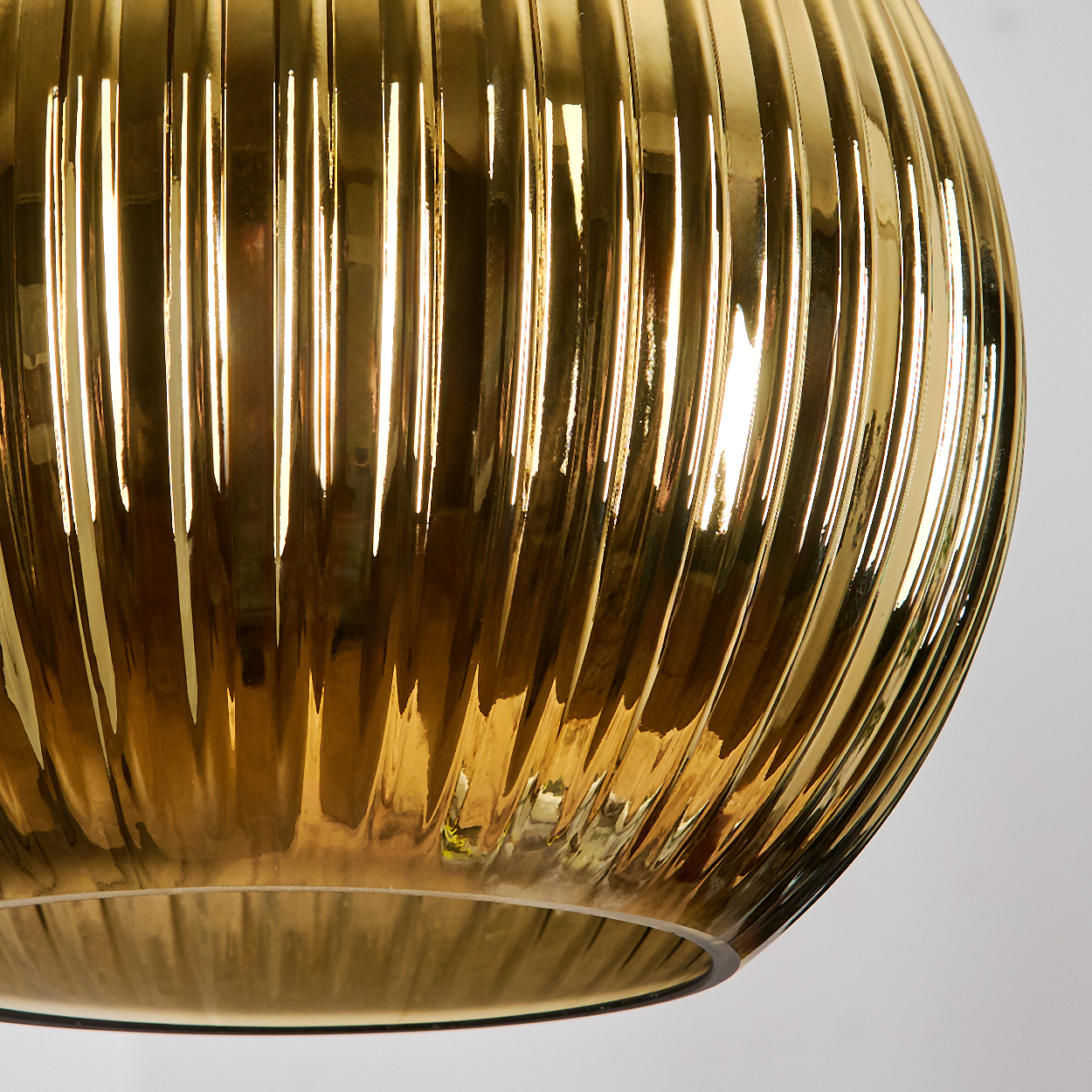Nahaufnahme einer goldenen, geriffelten Glasleuchte. Das Licht spiegelt sich auf der Glasoberfläche und erzeugt Glanzlichter und Schatten vor weißem Hintergrund.