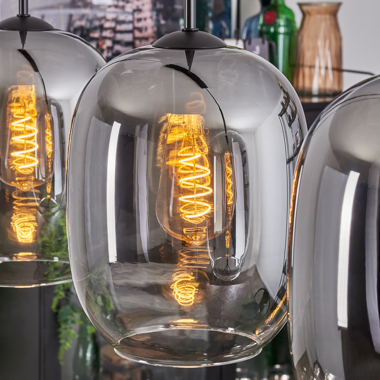 Drei Glaslampen mit leuchtenden Wendelbirnen. Lampen sind grau. Hintergrund hat Vasen und Dekoration.